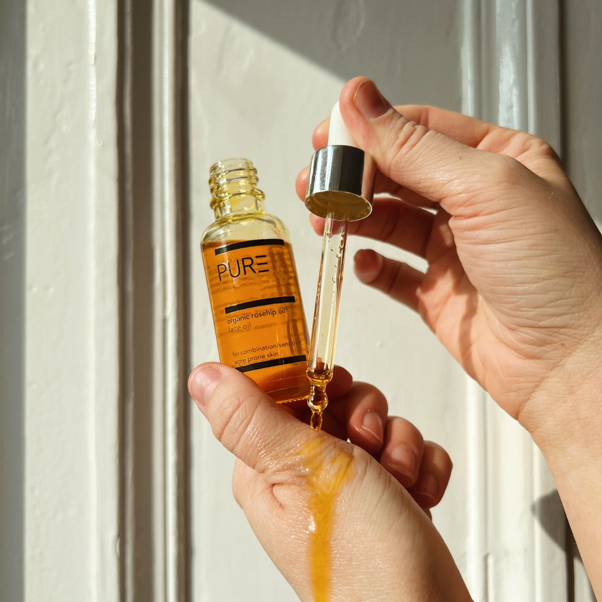 A hand holding a bottle of PURE Organic Cold-Pressed Rosehip Face Oil with a drop on the dropper
