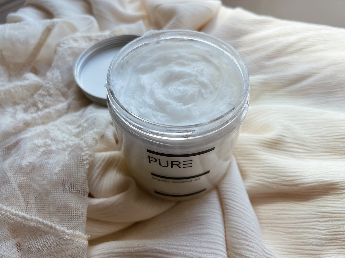 A jar of PURE Organic Virgin Coconut Oil with the lid off on soft white linen
